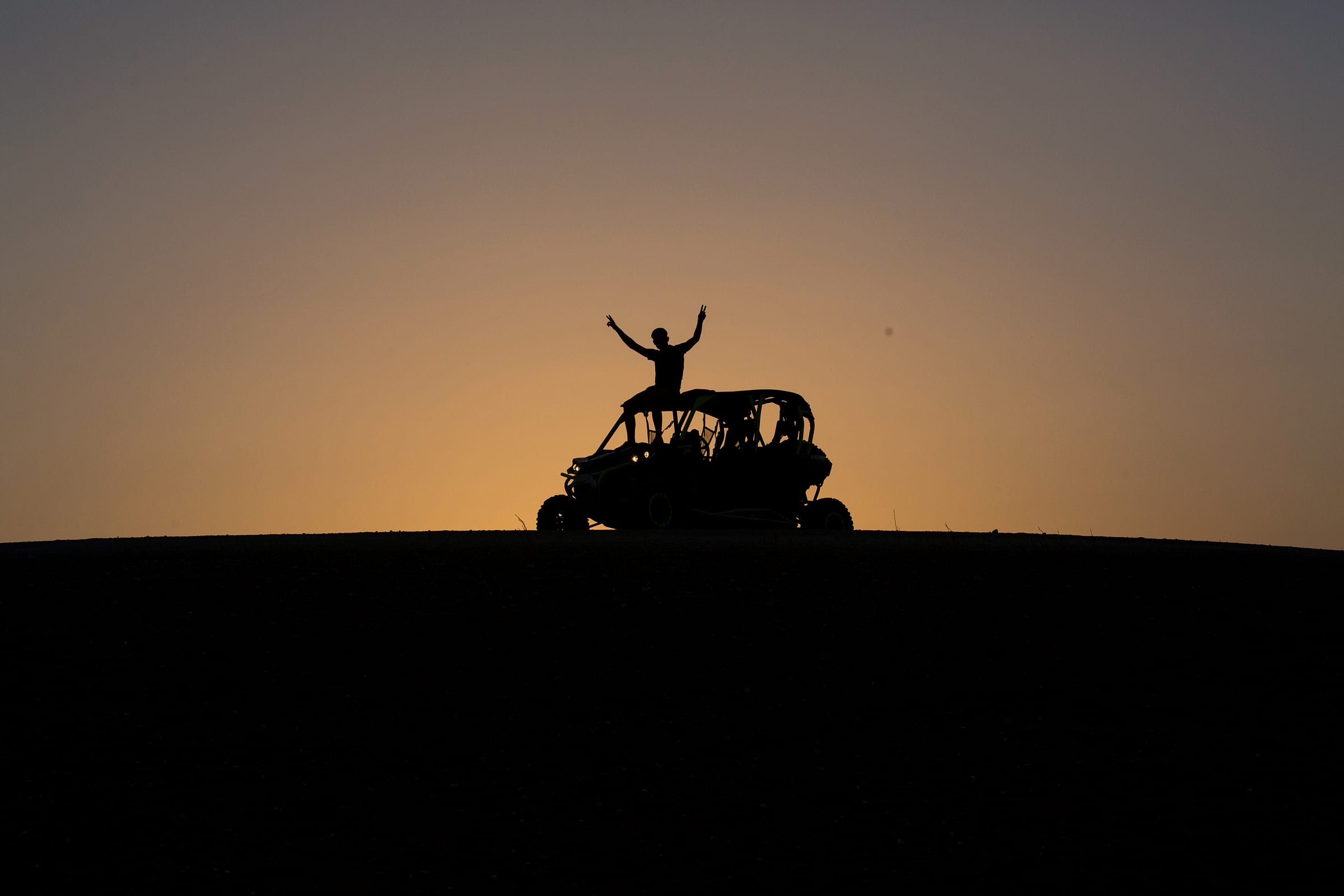 Silhouettes de pilotes bras levés sur leur buggy devant le ciel orangé du soir
