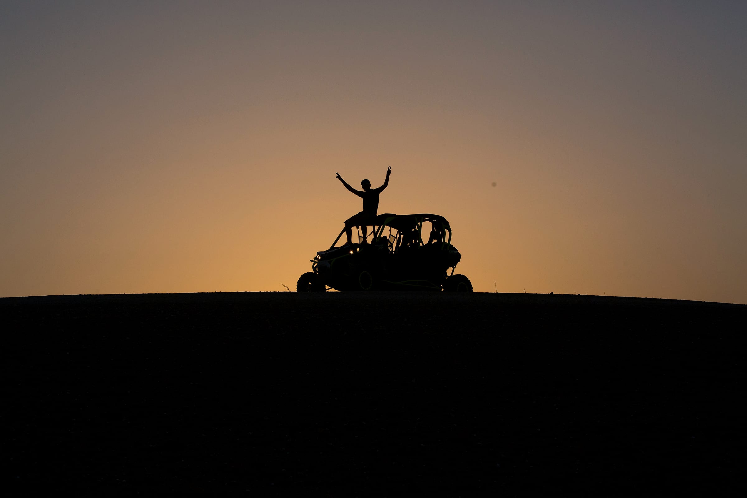 Silhouettes de pilotes bras levés sur leur buggy devant le ciel orangé du soir